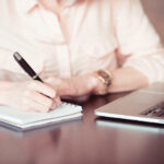Building Gender Discrimination Case: a woman writes in a notebook while sitting in front of a computer.