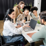 10 Signs of Disability Discrimination in the Workplace: a woman in a wheelchair sits at a table with other colleagues as they work together in the office.