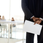 An African American businessman stands with his back to the viewer with a blurred background of coworkers