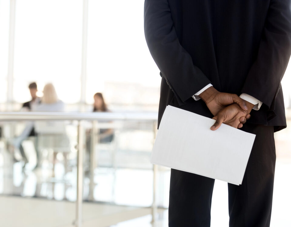An African American businessman stands with his back to the viewer with a blurred background of coworkers