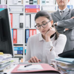 10 signs of a toxic work environment: Boss watching over his young employee and standing behind her, she is working and feeling oppressed