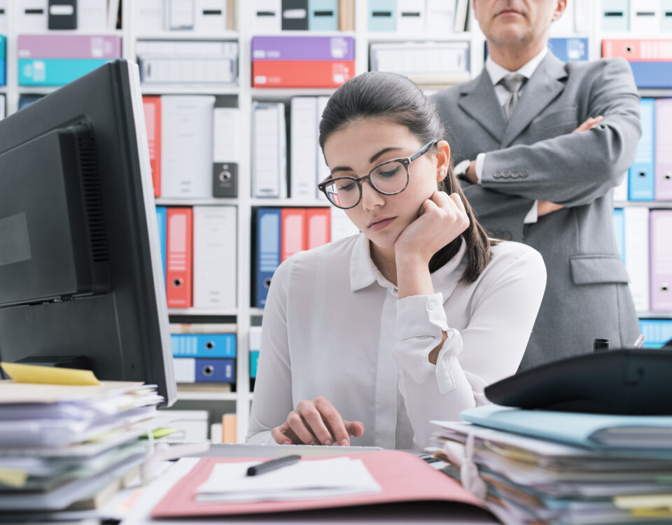10 signs of a toxic work environment: Boss watching over his young employee and standing behind her, she is working and feeling oppressed
