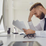 Workplace Retaliation: Upset busy businessman in a white shirt reads working documents sitting at a table with a computer in the office.
