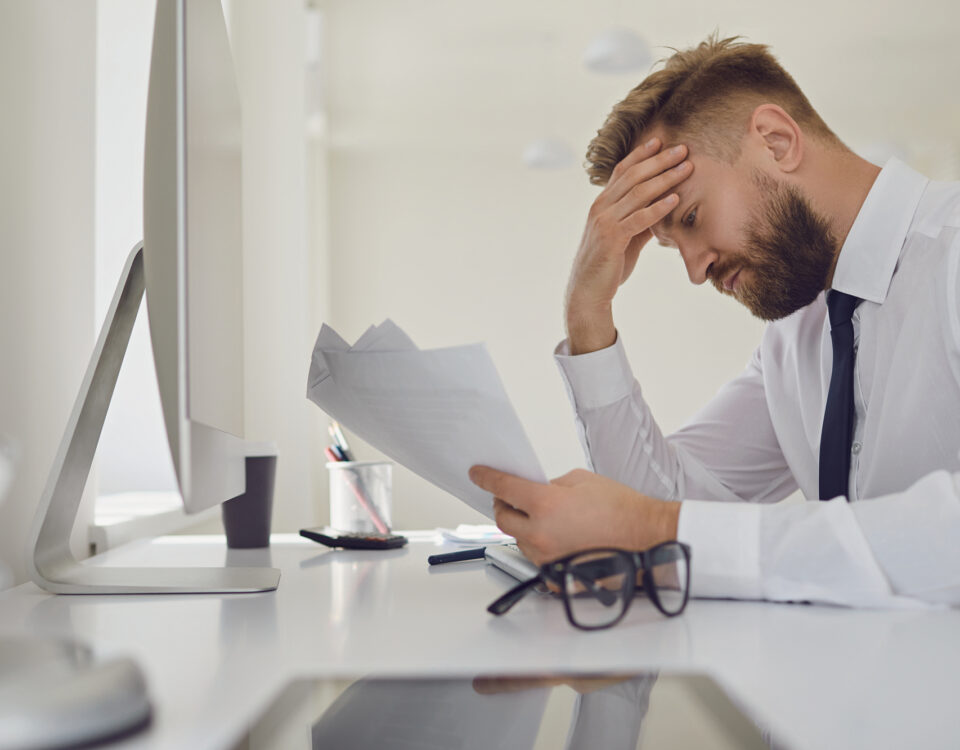 Workplace Retaliation: Upset busy businessman in a white shirt reads working documents sitting at a table with a computer in the office.