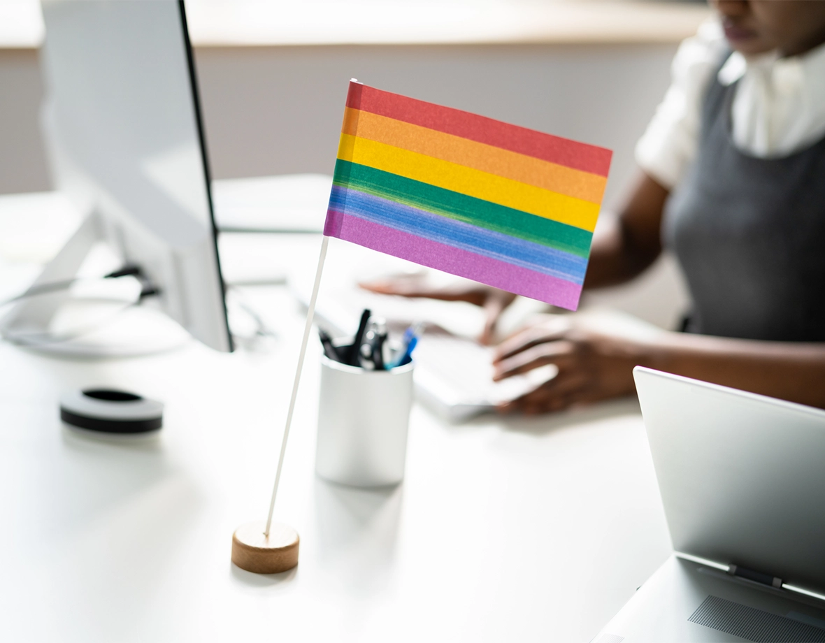 A mini LGBTQIA+ flag sits on an office desk filled with office supplies.